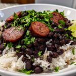 A close-up of a white bowl filled with white rice topped with seasoned black beans and rice, sliced sausage, and fresh cilantro, garnished with a lime wedge.