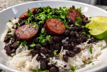 A close-up of a white bowl filled with white rice topped with seasoned black beans and rice, sliced sausage, and fresh cilantro, garnished with a lime wedge.