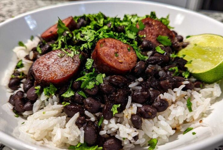 A close-up of a white bowl filled with white rice topped with seasoned black beans and rice, sliced sausage, and fresh cilantro, garnished with a lime wedge.