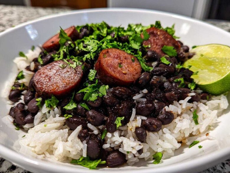 A close-up of a white bowl filled with white rice topped with seasoned black beans and rice, sliced sausage, and fresh cilantro, garnished with a lime wedge.