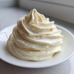 A tall, perfectly piped swirl of light, fluffy cream cheese frosting sitting on a small white saucer.
