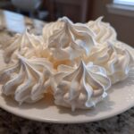 A close-up of several perfectly piped, white meringue cookies stacked on a small white plate.
