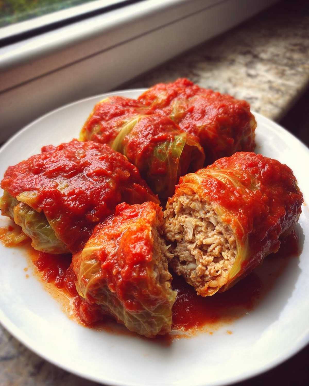 Four homemade cabbage rolls smothered in bright red tomato sauce, one cut open to show the savory meat and rice filling.