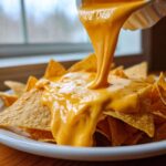 Thick, smooth nacho cheese sauce being poured over a pile of tortilla chips on a white plate.