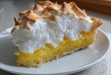 A close-up slice of lemon meringue pie showing a thick layer of bright yellow filling and towering, toasted meringue peaks.