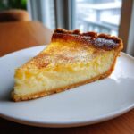 A close-up of a thick slice of creamy egg custard pie dusted with cinnamon on a white plate.