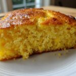 Close-up of a thick, moist slice of yellow skillet cornbread showing its crumb texture on a white plate.
