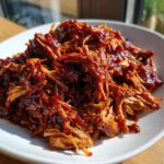 A close-up of tender, shredded Slow Cooker Pulled Pork heavily coated in rich, dark BBQ sauce, served on a white plate.