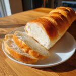 A freshly baked loaf of soft crusty french bread, partially sliced, resting on a white plate.