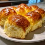 A cluster of freshly baked, soft dinner rolls with a shiny, golden-brown top, served on a white plate.