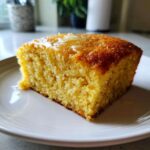 Close-up of a golden slice of sourdough cornbread topped with melting butter on a white plate.