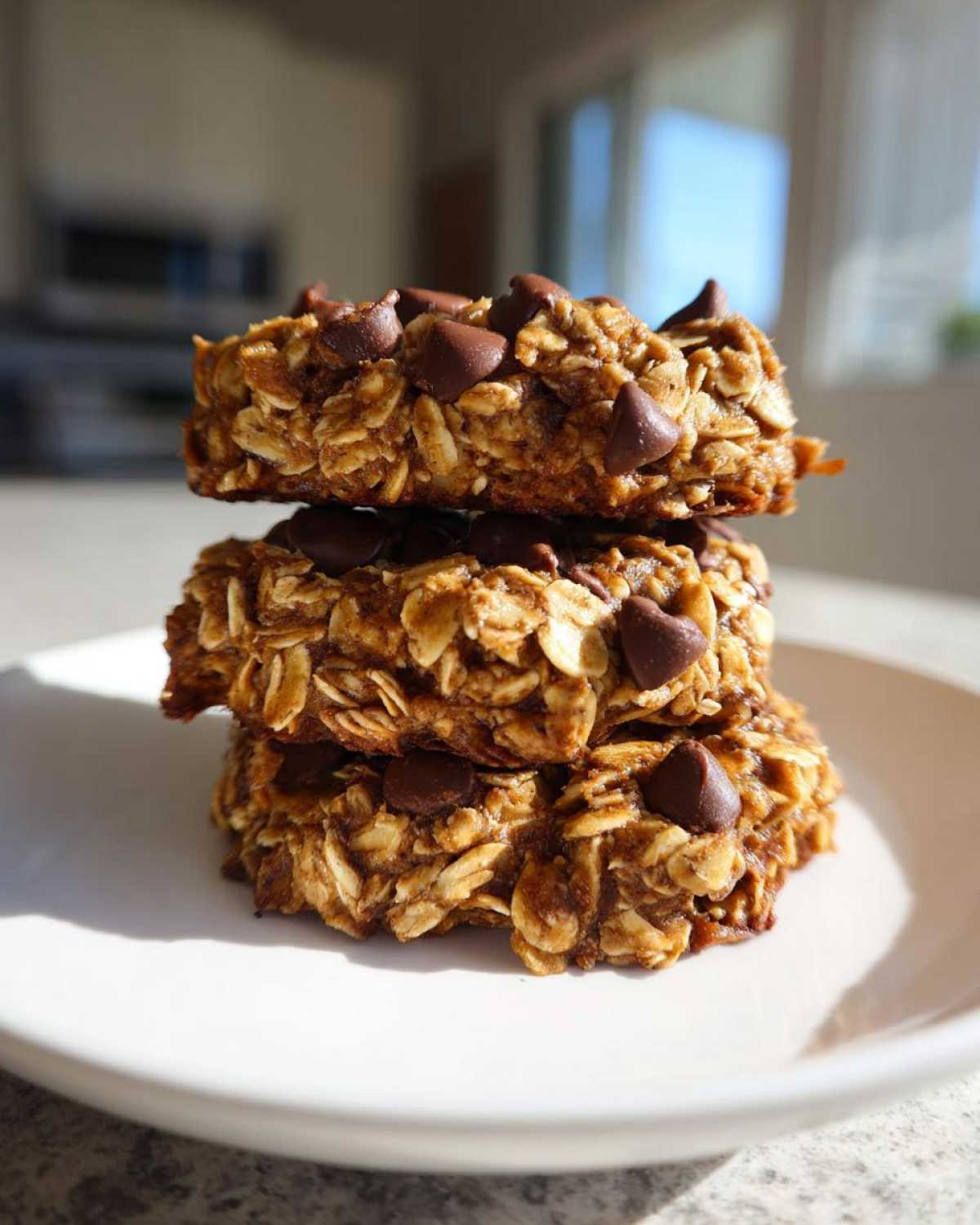 Three stacked, thick breakfast cookie treats made with oats and topped with chocolate chips, sitting on a white plate.