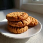 A stack of three golden brown peanut butter cookies featuring classic crisscross fork marks, served on a white plate.