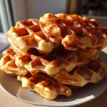 A tall stack of golden brown, freshly made high protein waffles piled on a small white plate near a window.