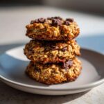 Three stacked, golden-brown breakfast cookie treats loaded with oats and chocolate chips on a light gray plate.