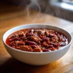 A close-up of a steaming white bowl filled with rich, red chili featuring ground meat and kidney beans, part of the best chili recipe.