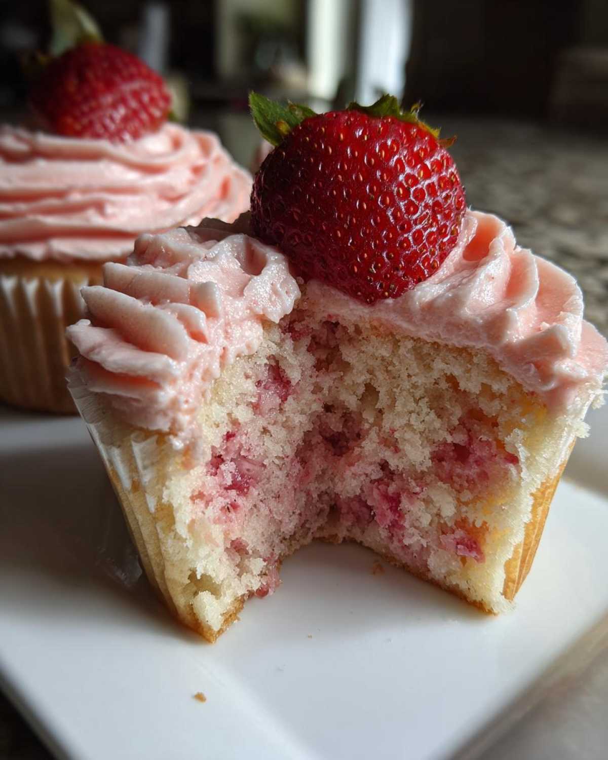 Close-up of a strawberry cupcake cut in half showing the moist cake and strawberry filling, topped with pink frosting and a fresh strawberry.
