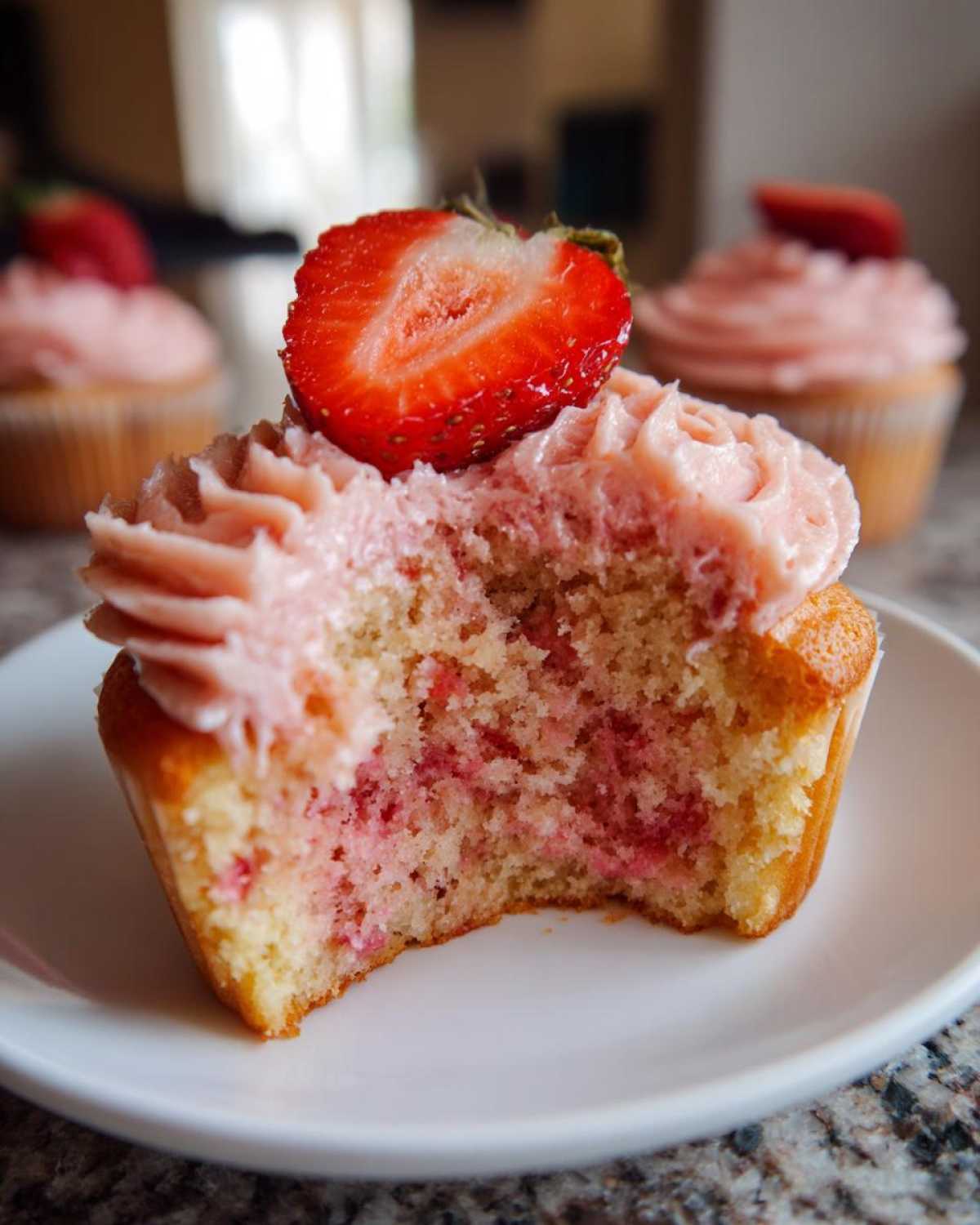 Close-up of a strawberry cupcake with a bite taken out, showing the moist cake and pink frosting topped with a fresh strawberry slice.