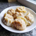 A white plate holding six freshly steamed Traditional Chinese Dumplings with slightly browned bottoms.