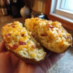 A person holding two halves of a twice baked potato stuffed with creamy filling, topped with melted cheddar cheese and bacon bits.