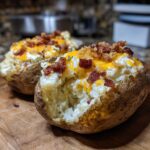 Two loaded twice baked potatoes topped with melted cheddar cheese, crispy bacon bits, and herbs on a wooden cutting board.