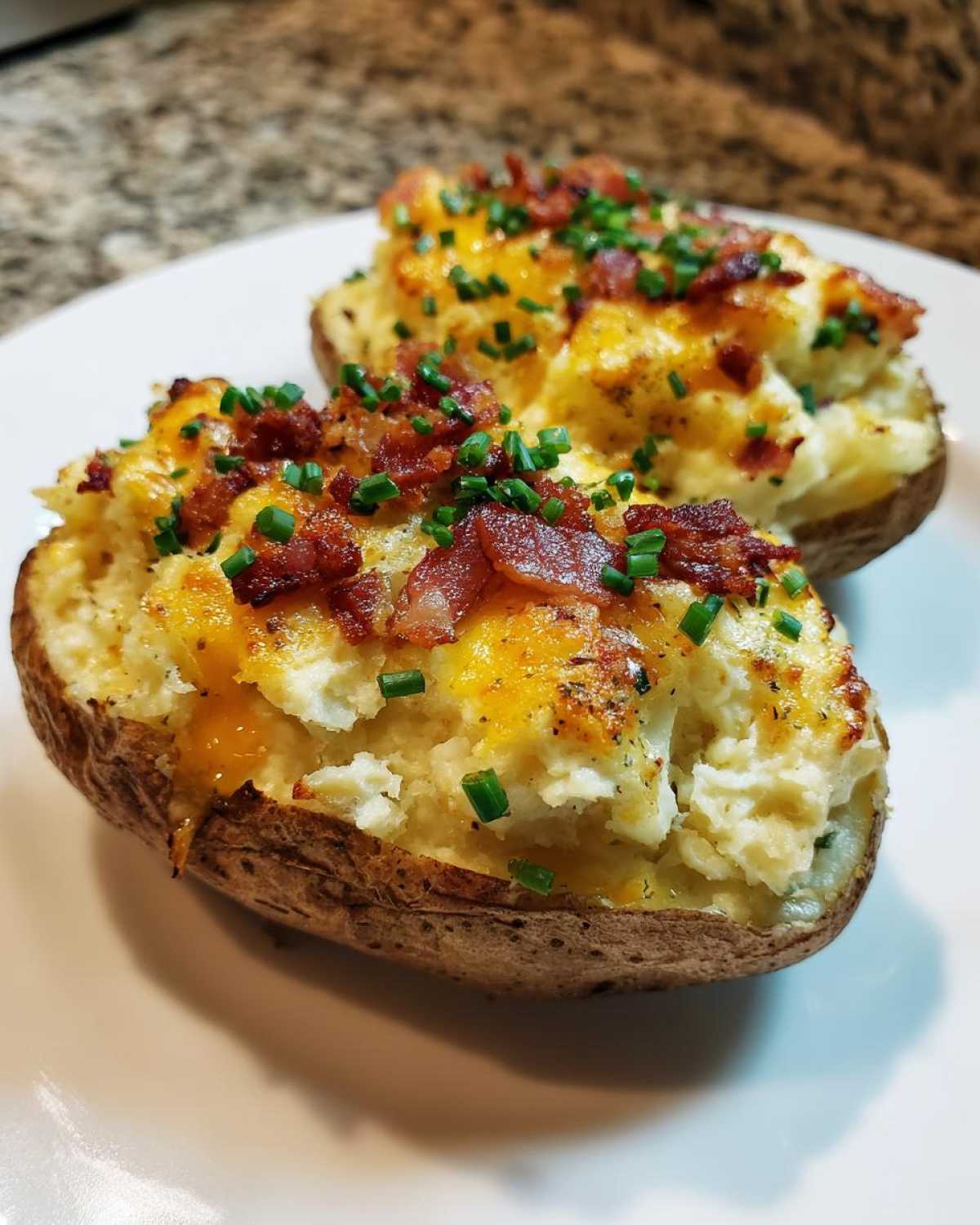 Two perfectly prepared twice-baked potatoes loaded with cheese, bacon bits, and fresh chives on a white plate.
