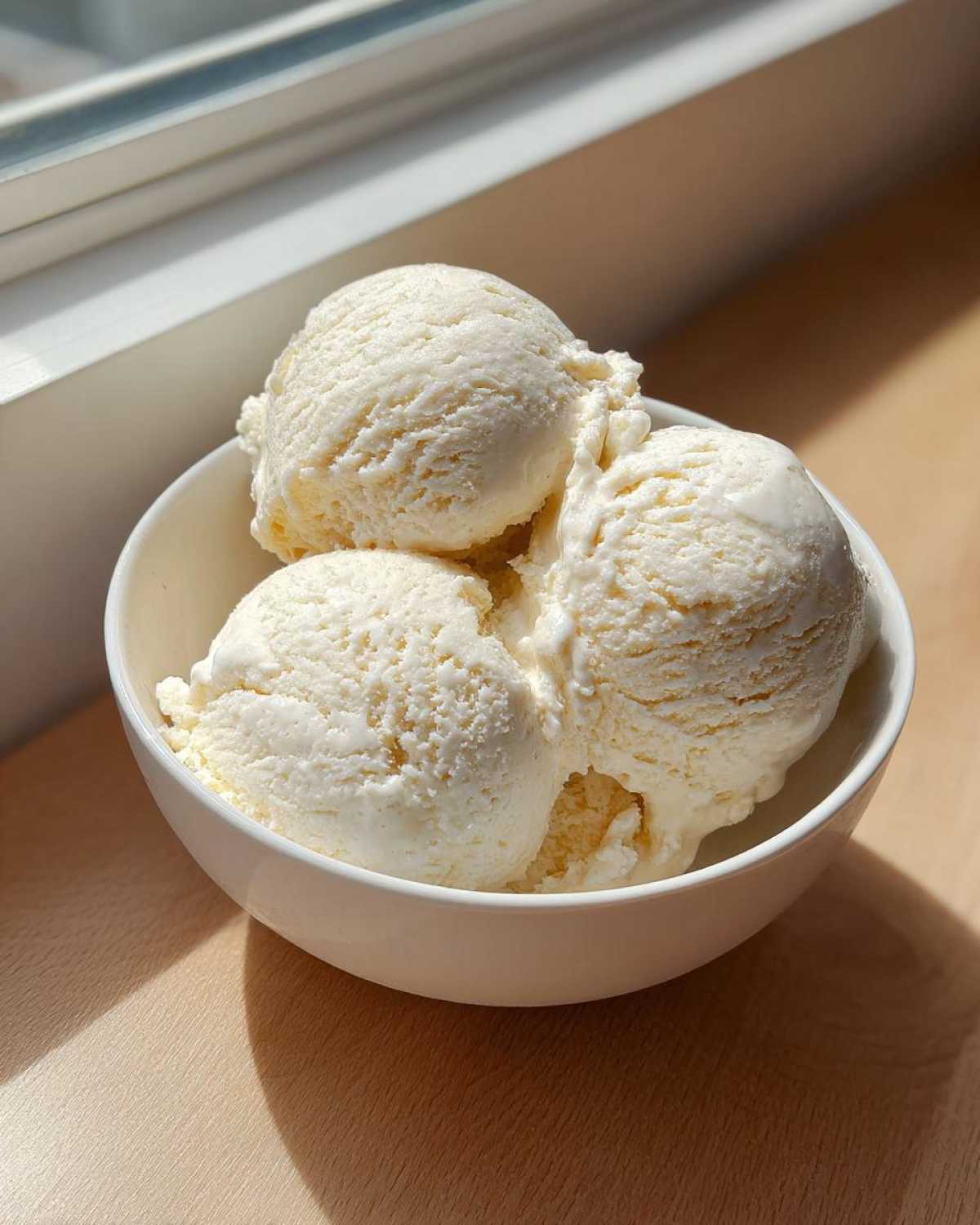 Three generous scoops of creamy vanilla protein ice cream served in a white bowl, brightly lit by sunlight.