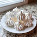 A pile of freshly baked, crisp white meringue cookies with piped swirls, served on a white plate.