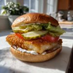 A close-up of an Air Fryer Crispy Fish Sandwich featuring a thick, golden fillet, pickles, tomato, lettuce, and tartar sauce on a brioche bun.