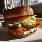 A close-up of an Air Fryer Crispy Fish Sandwich featuring a golden fillet, lettuce, pickles, tomato, and tartar sauce on a brioche bun.