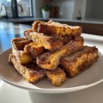 A tall stack of golden Air Fryer French Toast Sticks generously coated in sparkling cinnamon sugar on a white plate.