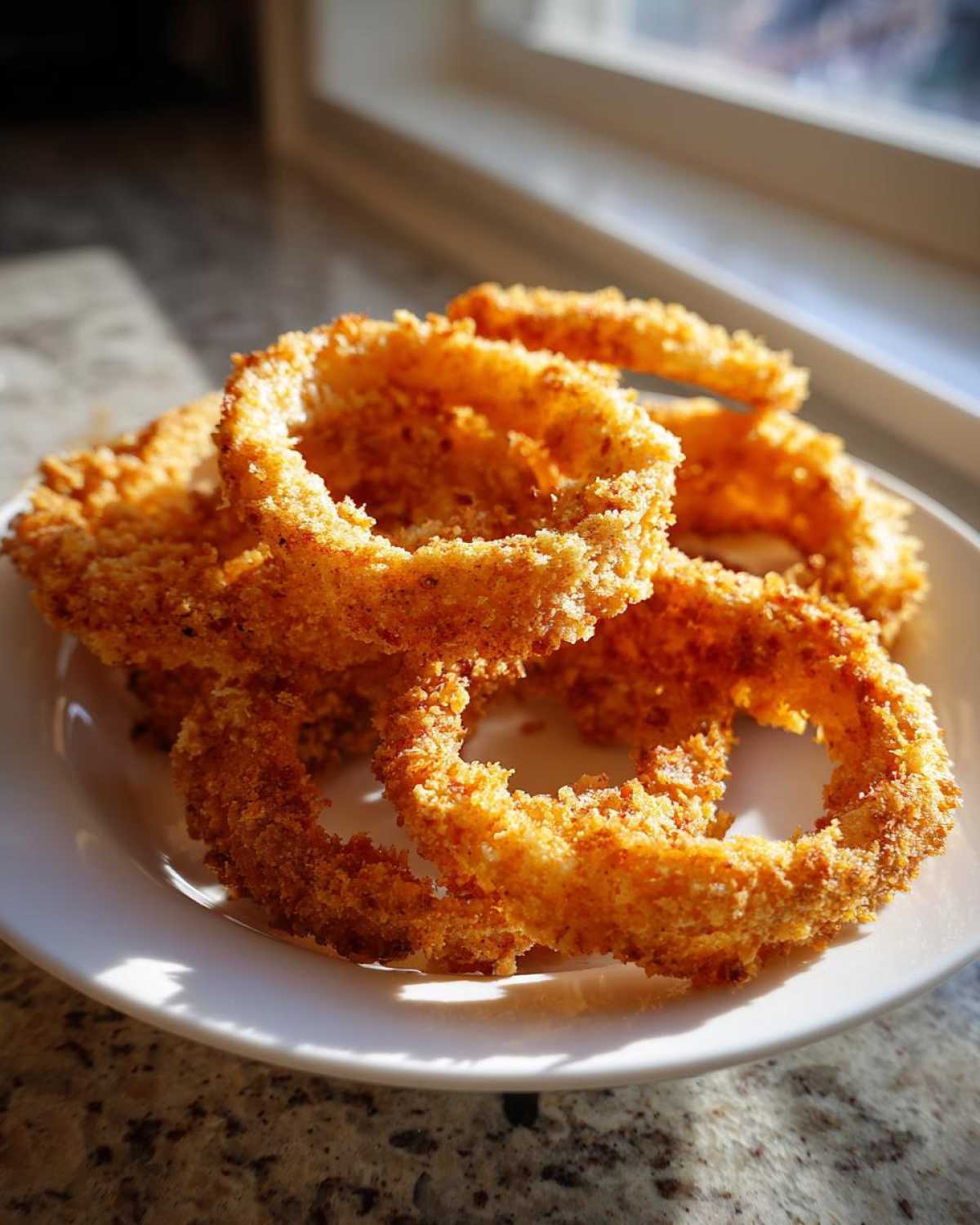 A close-up of several golden brown, crispy Air Fryer Onion Rings stacked on a white plate near a sunlit window.
