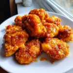 Close-up of crispy, glazed pieces of Air Fryer Orange Chicken piled on a white plate near a window.