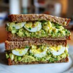 A tall, stacked Avocado Egg Salad Sandwich made with whole wheat bread and visible slices of hard-boiled egg.