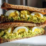 A tall stack of two halves of an Avocado Egg Salad Sandwich on toasted whole wheat bread, showing creamy green filling and sliced hard-boiled eggs.