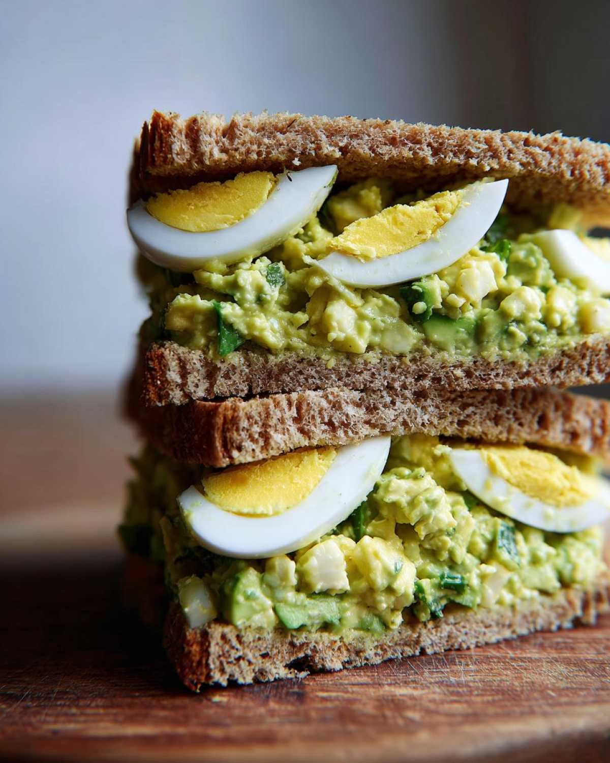 Close-up of a stacked Avocado Egg Salad Sandwich made with whole wheat bread and garnished with hard-boiled egg slices.
