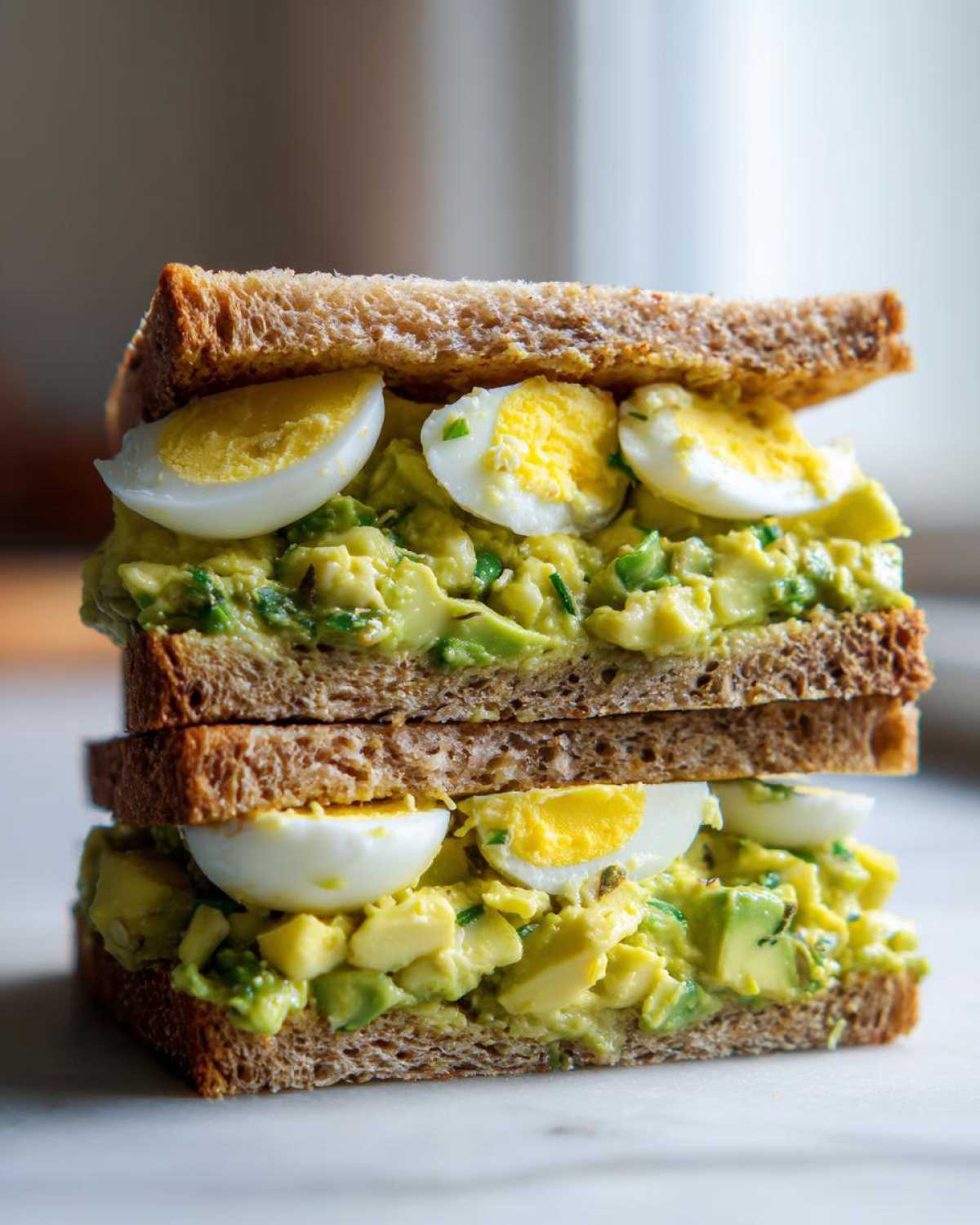 A tall, stacked Avocado Egg Salad Sandwich made with whole wheat bread and visible slices of hard-boiled egg.
