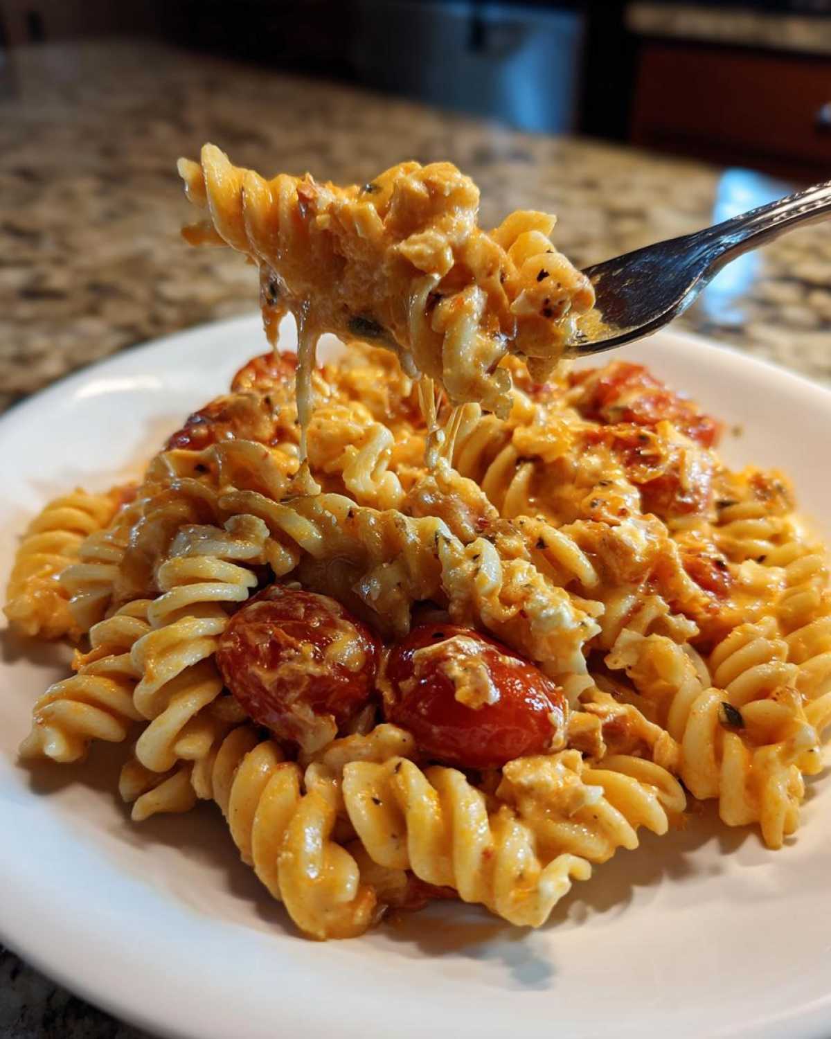 A fork lifting a cheesy, creamy portion of Baked Feta Pasta with Cherry Tomatoes from a white plate.