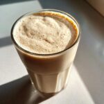 A tall glass filled with a creamy, frothy Banana Peanut Butter Smoothie, catching bright sunlight.
