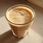 Close-up of a frothy Banana Peanut Butter Smoothie in a clear glass, sitting on a light countertop.