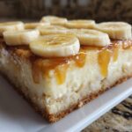 Close-up of a rich slice of Banana Pudding Cheesecake Bars topped with caramel sauce and fresh banana slices.