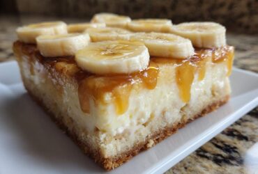 Close-up of a rich slice of Banana Pudding Cheesecake Bars topped with caramel sauce and fresh banana slices.