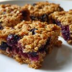 Close-up of several golden-brown Blueberry Baked Oatmeal Squares with visible baked blueberries on a white plate.