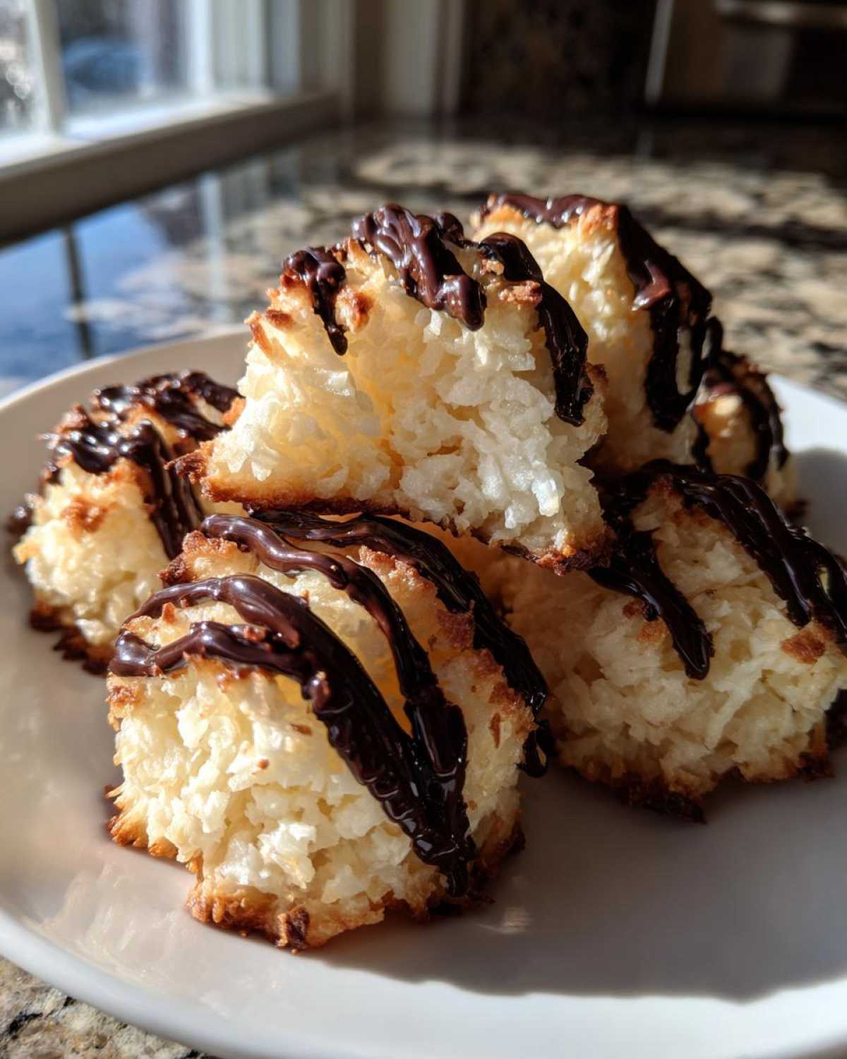 A stack of golden Coconut Macaroons with Dark Chocolate Drizzle resting on a white plate in bright sunlight.