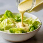 Thick, creamy Copycat Olive Garden Salad Dressing being poured from a jar onto crisp romaine lettuce in a white bowl.