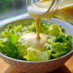 Pouring creamy Copycat Olive Garden Salad Dressing over crisp green lettuce in a white bowl.