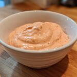 A white bowl filled with creamy, orange-tinted Copycat Raising Cane’s Sauce, resting on a wooden cutting board.