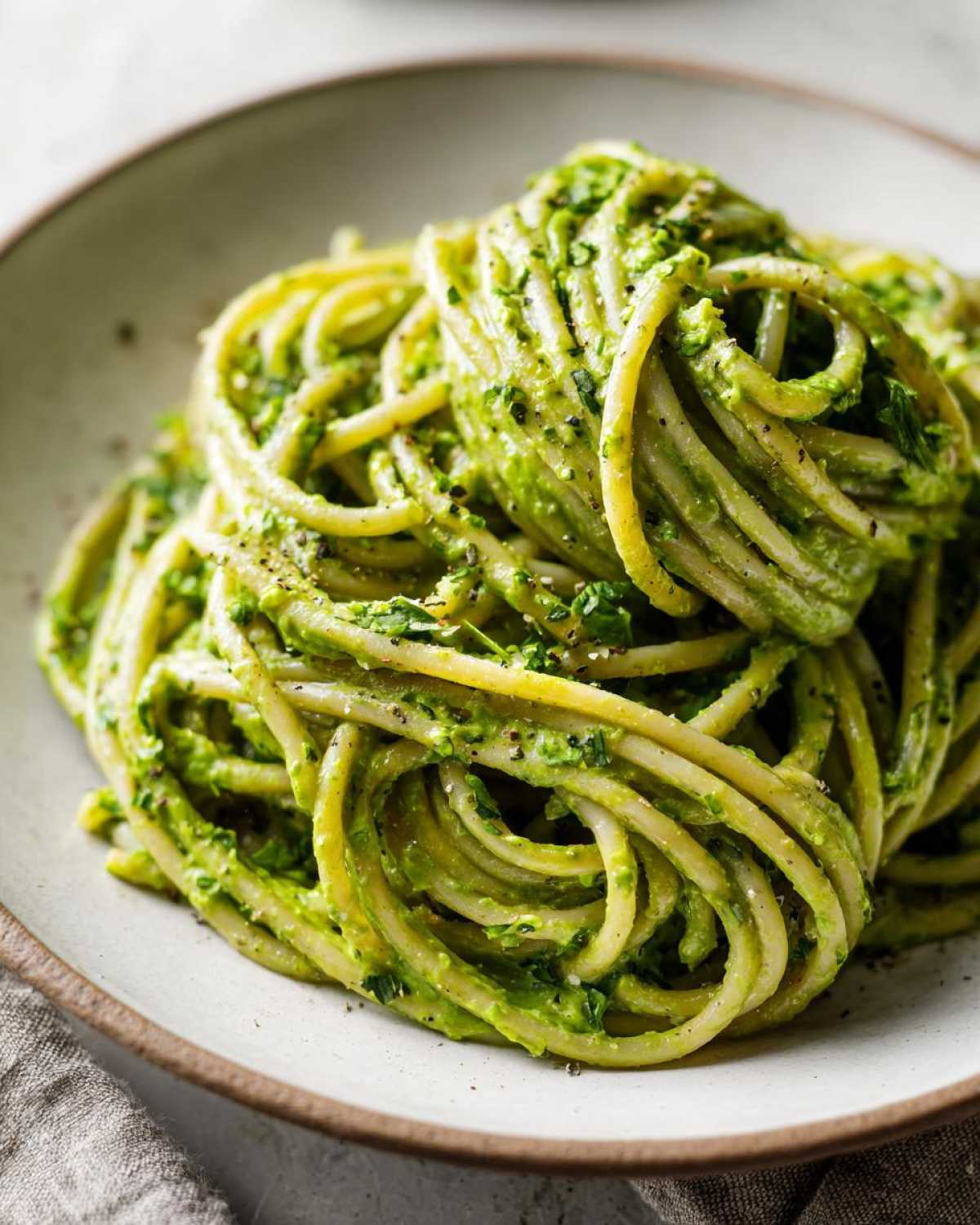 A close-up shot of perfectly twirled spaghetti coated in vibrant green Creamy Avocado Pasta sauce, seasoned with black pepper.