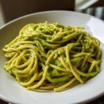 A close-up shot of spaghetti generously coated in bright green, creamy avocado pasta sauce served in a white bowl.