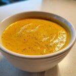 Close-up of a white bowl filled with bright orange Creamy Coconut Lentil Soup, topped with dark spices.